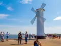 Biała latarnia morska w kształcie wiatraka na plaży w Świnoujściu, z turystami spacerującymi po molo.