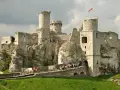 Ruiny zamku Ogrodzieniec na skale, z polską flagą na wieży i grupą turystów na moście.
