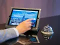 Dłoń dotyka ekranu tabletu, rozpoczynając proces zameldowania w hotelu. Obok stoi dzwonek hotelowy.
