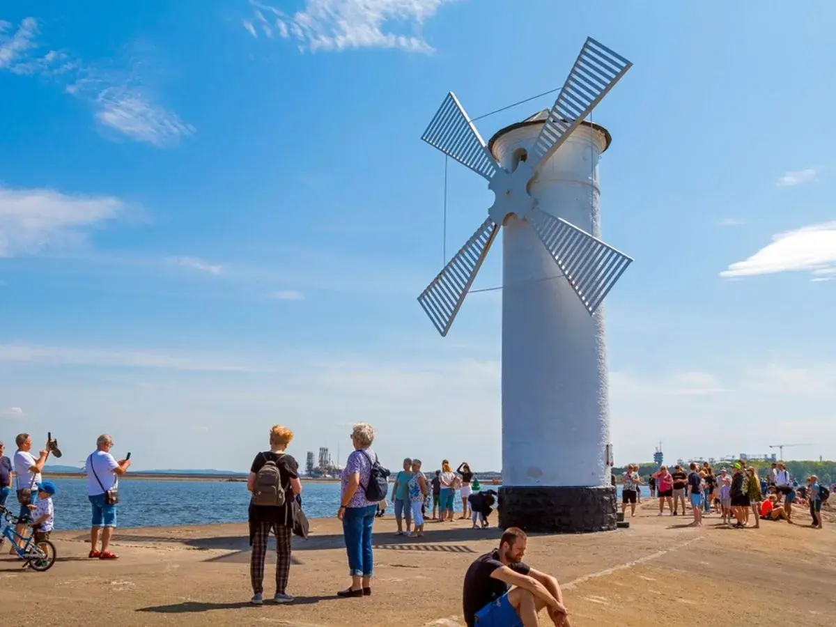 Biała latarnia morska w kształcie wiatraka na plaży w Świnoujściu, z turystami spacerującymi po molo.