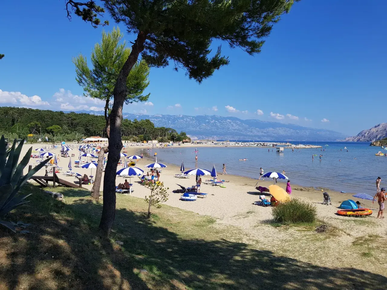 Słoneczny dzień na plaży, pełnej parasoli i ludzi. Woda jest spokojna, a w oddali widać góry. To prawdziwy raj na ziemi.
