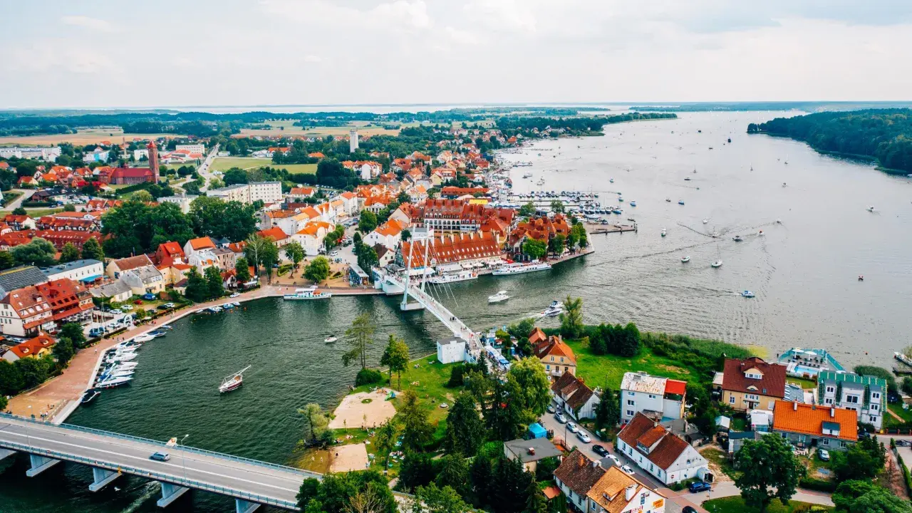Malownicze miasta na Mazurach z widokiem na jezioro i liczne łodzie. Biały most łączy brzegi, a czerwone dachy domów tworzą urokliwy krajobraz.