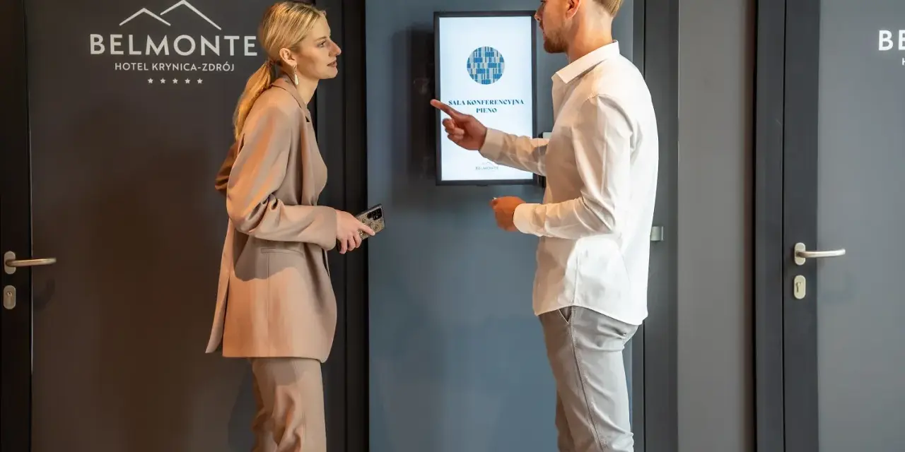 Elegancka para dyskutuje przy hotelowym ekranie. Ona w beżowym garniturze, on w białej koszuli. Idealny strój na śniadanie w hotelu.