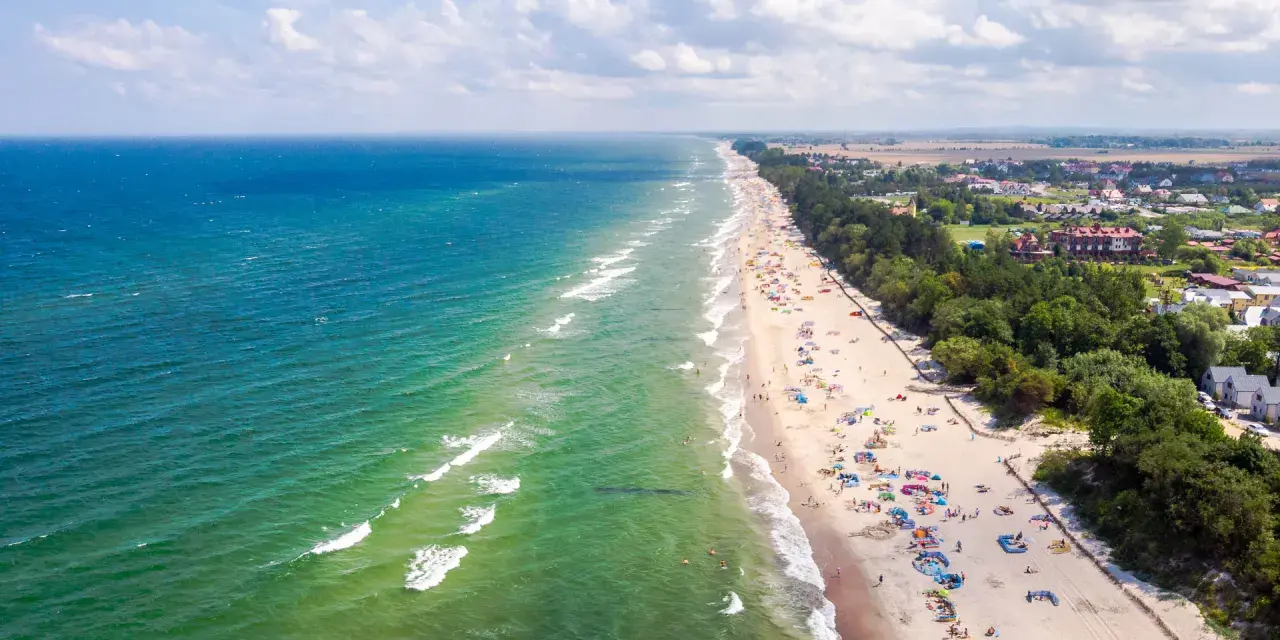 Słoneczny dzień na plaży w Sarbinowie nad morzem. Tłumy turystów odpoczywają na piasku, kąpią się w morzu i spacerują wzdłuż brzegu.