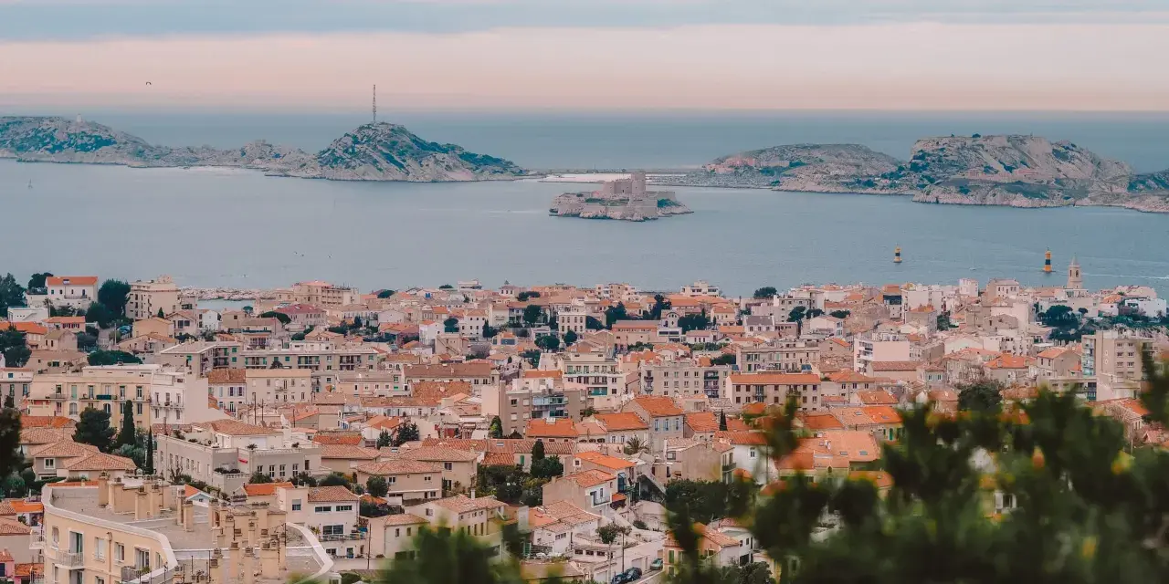 Widok na miasto z dachami i morzem w tle, z widocznym zamkiem na wyspie. To piękne **Marseille**.