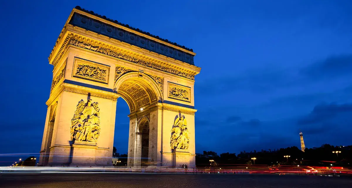 Łuk Triumfalny nocą, z widoczną w oddali Wieżą Eiffla. Światła samochodów tworzą smugi na tle **Champs-Élysées**.