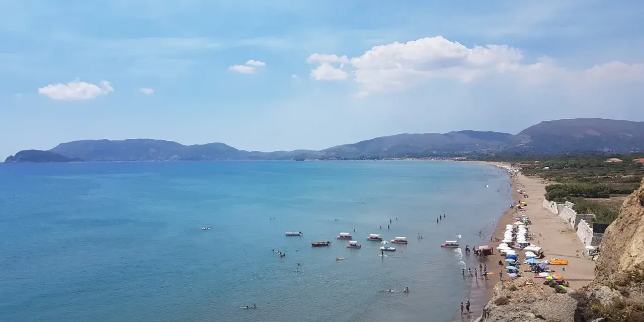 Słoneczny dzień na plaży Kalamaki. Ludzie pływają, opalają się i relaksują na piasku. Woda jest spokojna i turkusowa.