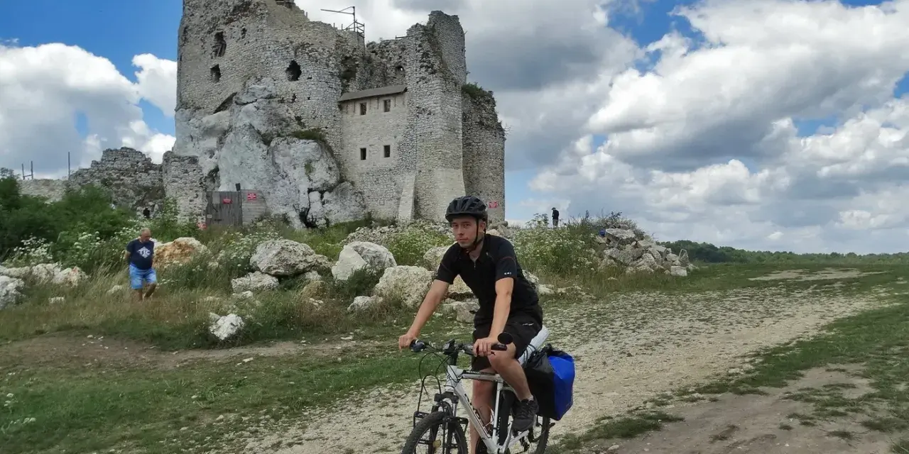 Młody rowerzysta przemierza malownicze **Jura Krakowsko-Częstochowska szlaki**, mijając ruiny zamku na tle błękitnego, chmurnego nieba.