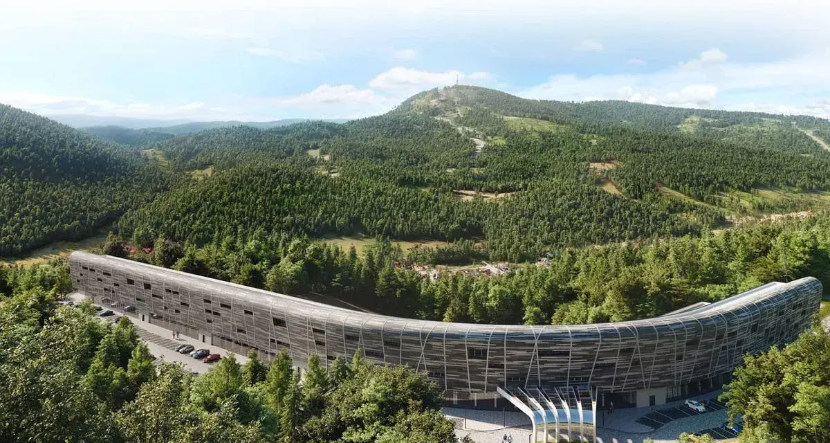 Nowy hotel w górach, o futurystycznym, zakrzywionym kształcie, otoczony gęstym lasem iglastym. W tle widać łagodne, zalesione wzgórza.