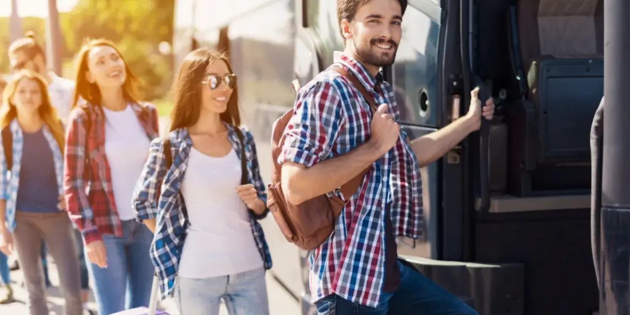 Grupa młodych ludzi z plecakami i walizką wsiada do autobusu, gotowa na wycieczki fakultatywne.