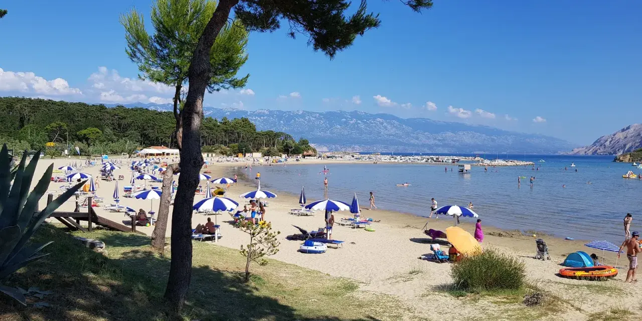 Słoneczny dzień na plaży, pełnej parasoli i ludzi. Woda jest spokojna, a w oddali widać góry. To prawdziwy raj na ziemi.