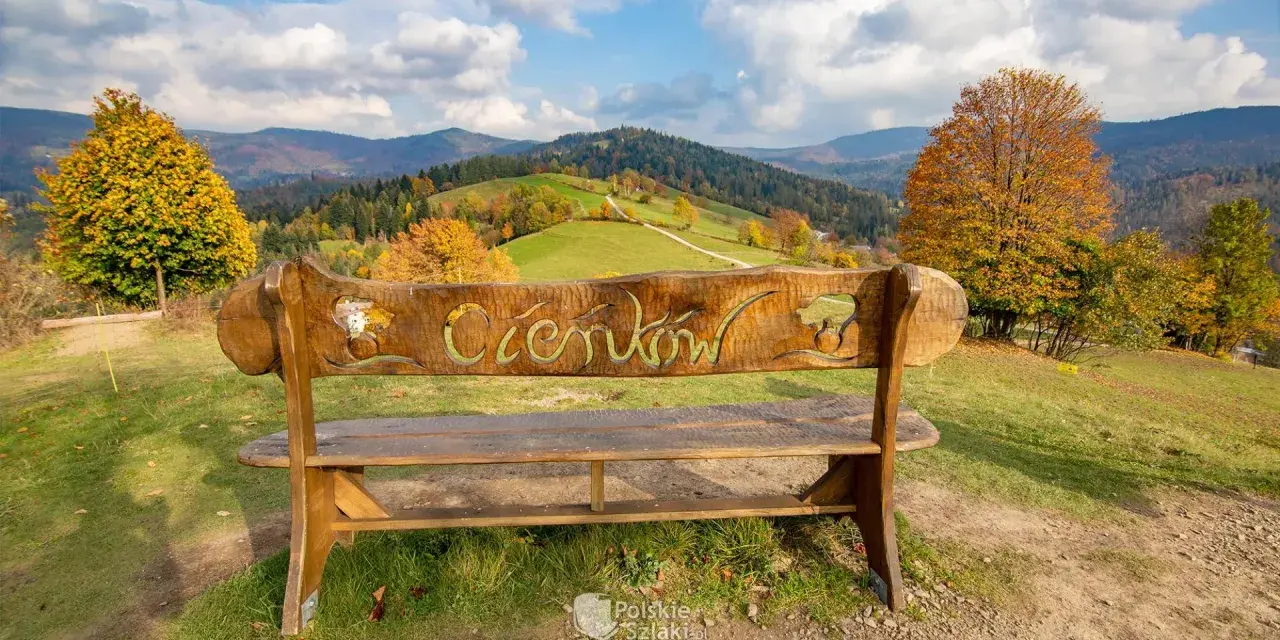 Drewniana ławka z napisem "Ciechocinek" na tle jesiennych gór i zielonych wzgórz. Widok na Beskid Sądecki.