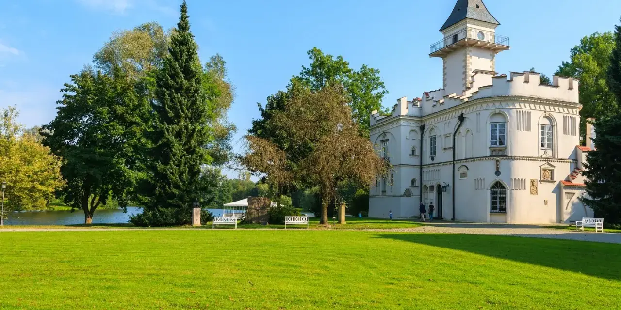 Biały pałac z wieżą, otoczony zielenią, nad jeziorem. Jeden z urokliwych zamków na Mazowszu.