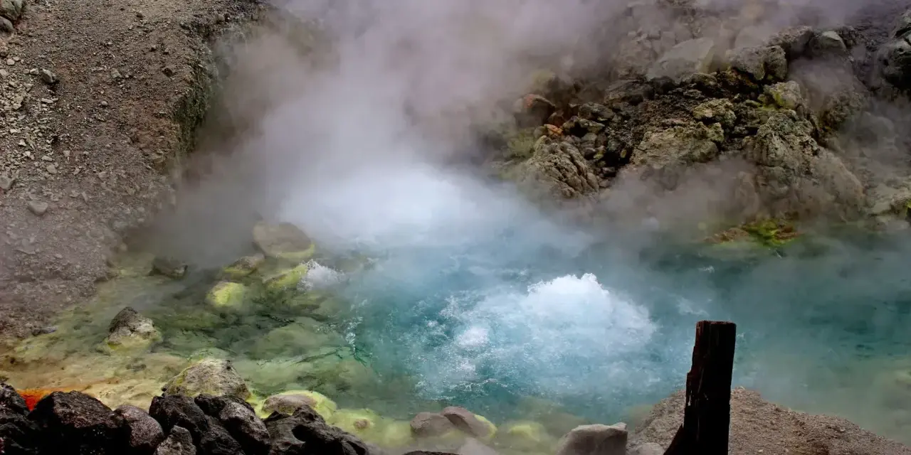 Gorący, bulgoczący onsen otoczony skalistym, parującym krajobrazem. Woda ma turkusowy odcień, a brzegi pokryte są żółtymi osadami.