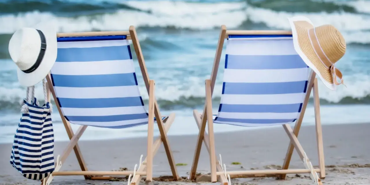 Dwa leżaki na plaży, gotowe na idealny weekend nad morzem. W tle fale, a obok kapelusze i torba plażowa.