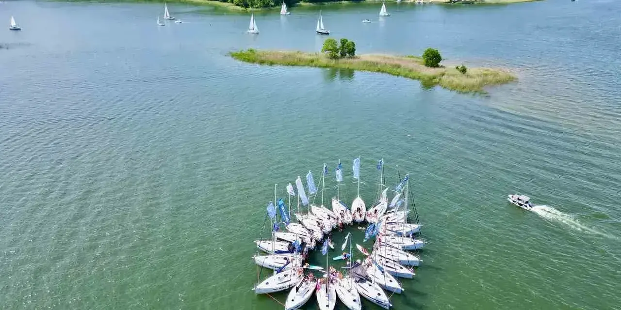 Żaglówki tworzą okrąg na spokojnym **lago**, otoczone zielonym lasem.