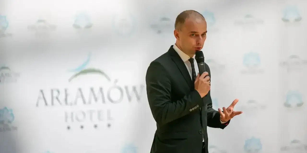 Mężczyzna w garniturze przemawia na tle logo Hotel Arłamów. Prawdopodobnie właściciel hotelu.