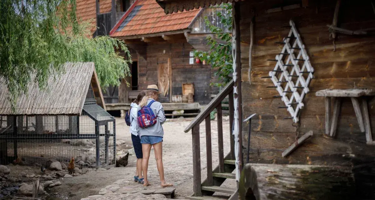 Dwie dziewczyny zwiedzają skansen, który może być częścią oferty hotelu bon turystyczny. W tle drewniane chaty i kurnik.