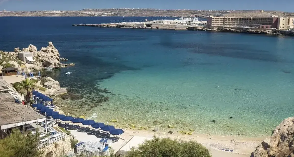 Rajska zatoka na Malcie z turkusową wodą, piaszczystą plażą i rzędem niebieskich parasoli. W oddali widać hotel i port.