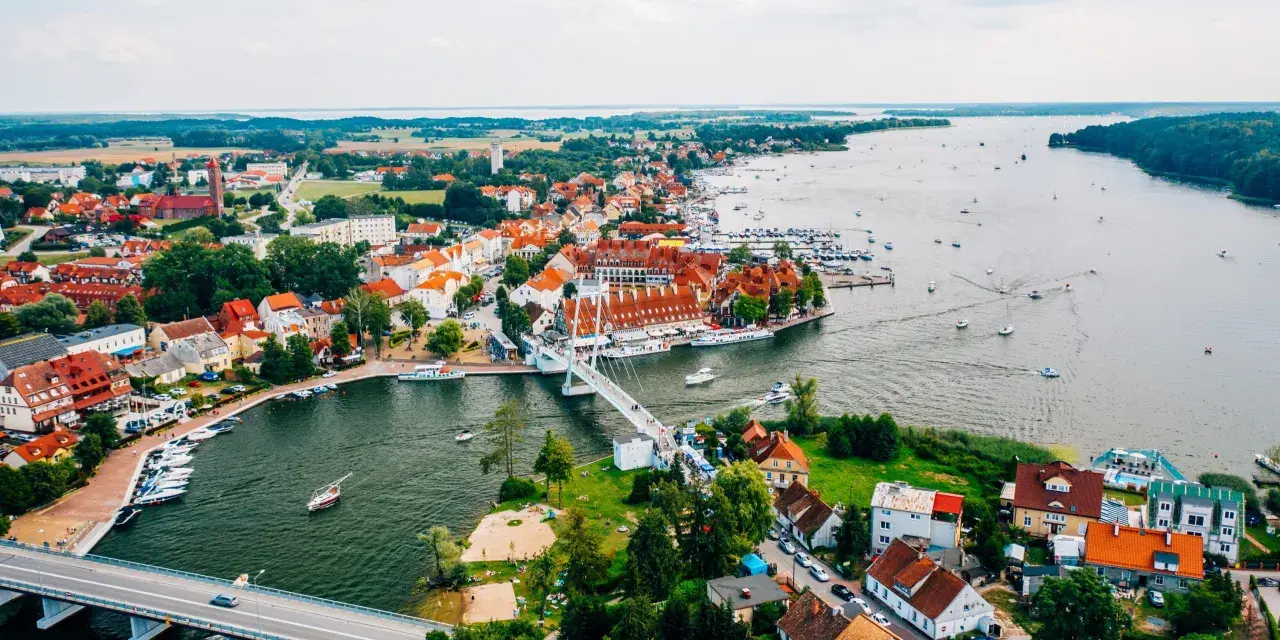 Malownicze miasta na Mazurach z widokiem na jezioro i liczne łodzie. Biały most łączy brzegi, a czerwone dachy domów tworzą urokliwy krajobraz.