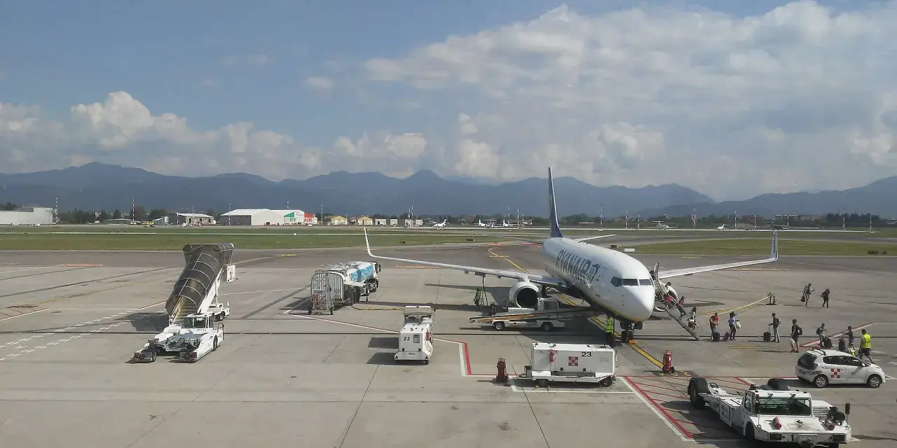 Samolot Ryanair na płycie lotniska Bergamo. W tle góry, na pierwszym planie schody, pojazdy obsługi naziemnej i pasażerowie.