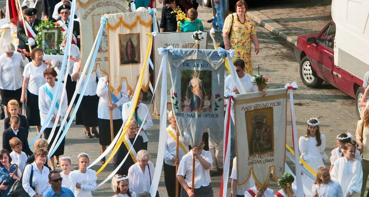 Procesja z banerami religijnymi, w tym z napisem "Boże Ciało 26". Ludzie niosą obrazy i kwiaty.