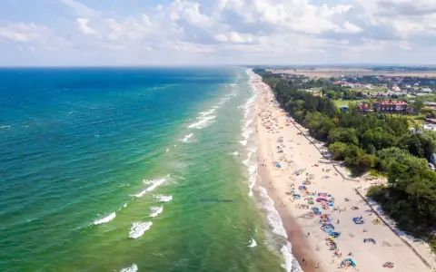 Słoneczny dzień na plaży w Sarbinowie nad morzem. Tłumy turystów odpoczywają na piasku, kąpią się w morzu i spacerują wzdłuż brzegu.