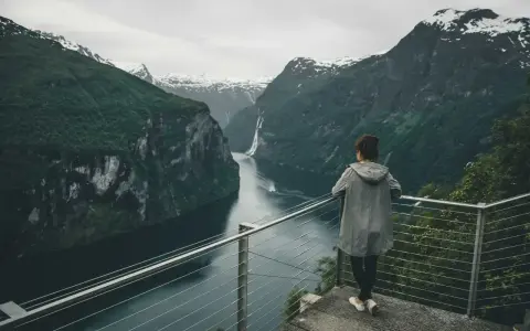Kobieta podziwia majestatyczne fiordy Norwegii. W oddali widać wodospad i łódź.