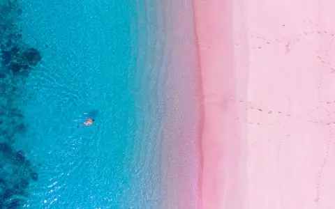Widok z lotu ptaka na turkusowe morze i różową plażę w Grecji. Ktoś pływa w krystalicznie czystej wodzie.