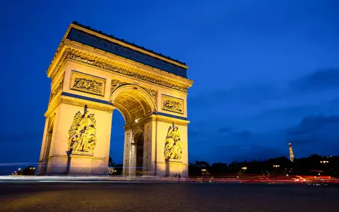 Łuk Triumfalny nocą, z widoczną w oddali Wieżą Eiffla. Światła samochodów tworzą smugi na tle **Champs-Élysées**.