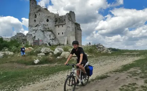 Młody rowerzysta przemierza malownicze **Jura Krakowsko-Częstochowska szlaki**, mijając ruiny zamku na tle błękitnego, chmurnego nieba.
