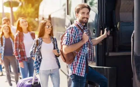 Grupa młodych ludzi z plecakami i walizką wsiada do autobusu, gotowa na wycieczki fakultatywne.