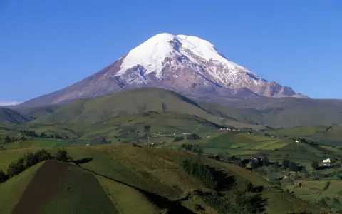 Pokryty śniegiem wulkan Chimborazo, jeden z najwyższych gór świata, góruje nad zielonymi, pofałdowanymi wzgórzami z polami uprawnymi.