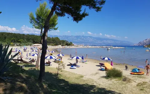 Słoneczny dzień na plaży, pełnej parasoli i ludzi. Woda jest spokojna, a w oddali widać góry. To prawdziwy raj na ziemi.