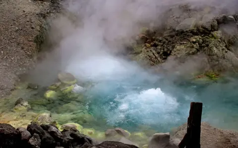 Gorący, bulgoczący onsen otoczony skalistym, parującym krajobrazem. Woda ma turkusowy odcień, a brzegi pokryte są żółtymi osadami.
