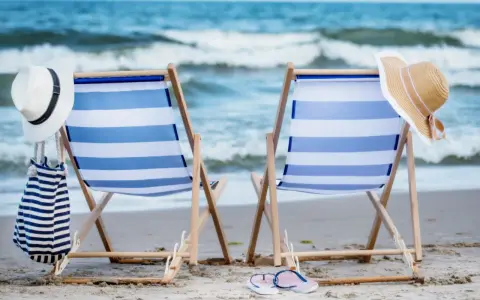 Dwa leżaki na plaży, gotowe na idealny weekend nad morzem. W tle fale, a obok kapelusze i torba plażowa.