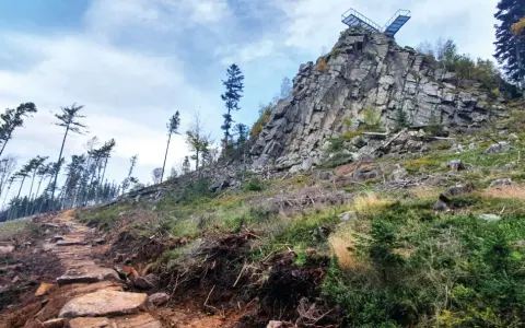Kamienista ścieżka prowadzi ku skalistemu szczytowi z platformą widokową. W oddali widać las, a krajobraz przypomina trojak Lądek Zdrój.
