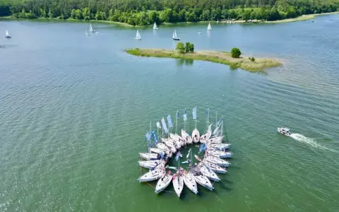 Żaglówki tworzą okrąg na spokojnym **lago**, otoczone zielonym lasem.