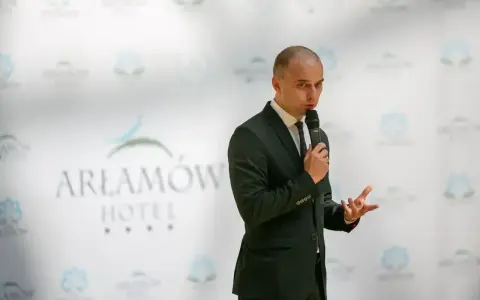 Mężczyzna w garniturze przemawia na tle logo Hotel Arłamów. Prawdopodobnie właściciel hotelu.