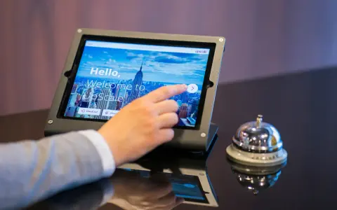 Dłoń dotyka ekranu tabletu, rozpoczynając proces zameldowania w hotelu. Obok stoi dzwonek hotelowy.