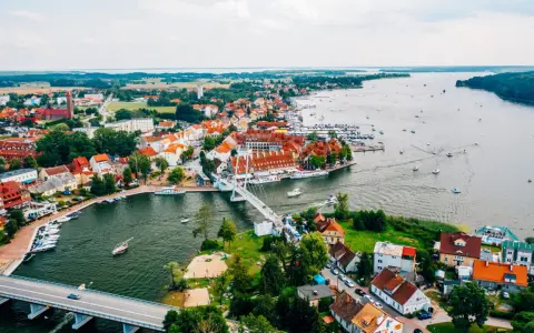 Malownicze miasta na Mazurach z widokiem na jezioro i liczne łodzie. Biały most łączy brzegi, a czerwone dachy domów tworzą urokliwy krajobraz.