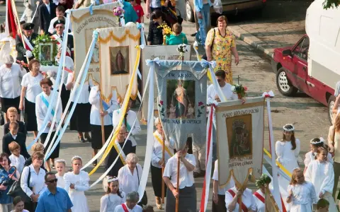 Procesja z banerami religijnymi, w tym z napisem "Boże Ciało 26". Ludzie niosą obrazy i kwiaty.
