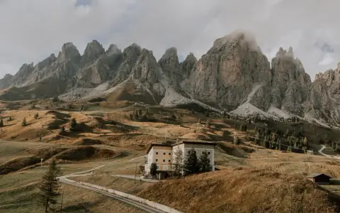 Malownicze **Val di Funes** z górami w tle, hotel na wzgórzu i kręta droga.