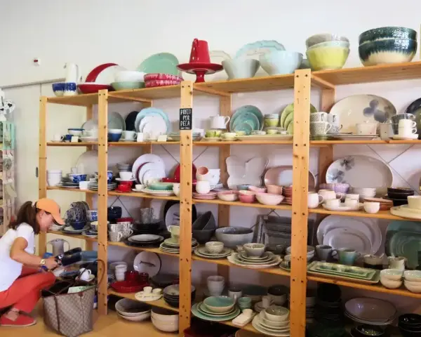 Kobieta wybiera ceramiczne naczynia, idealne pamiątki, które warto co przywieźć z Portugalii. Kolorowe talerze, miski i filiżanki na drewnianych półkach.