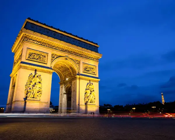Łuk Triumfalny nocą, z widoczną w oddali Wieżą Eiffla. Światła samochodów tworzą smugi na tle **Champs-Élysées**.