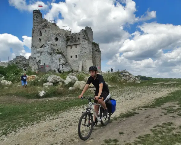 Młody rowerzysta przemierza malownicze **Jura Krakowsko-Częstochowska szlaki**, mijając ruiny zamku na tle błękitnego, chmurnego nieba.