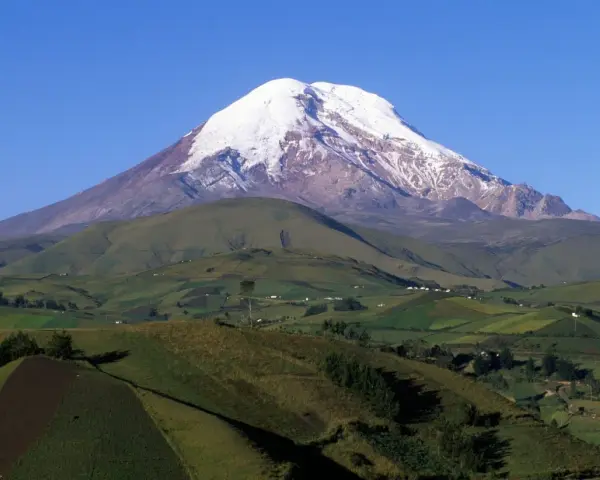 Pokryty śniegiem wulkan Chimborazo, jeden z najwyższych gór świata, góruje nad zielonymi, pofałdowanymi wzgórzami z polami uprawnymi.