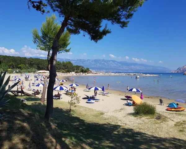 Słoneczny dzień na plaży, pełnej parasoli i ludzi. Woda jest spokojna, a w oddali widać góry. To prawdziwy raj na ziemi.