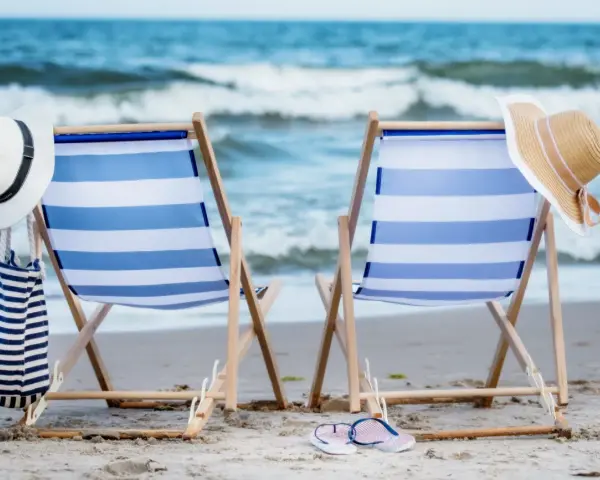 Dwa leżaki na plaży, gotowe na idealny weekend nad morzem. W tle fale, a obok kapelusze i torba plażowa.