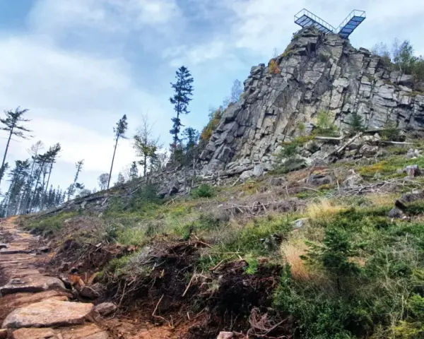 Kamienista ścieżka prowadzi ku skalistemu szczytowi z platformą widokową. W oddali widać las, a krajobraz przypomina trojak Lądek Zdrój.